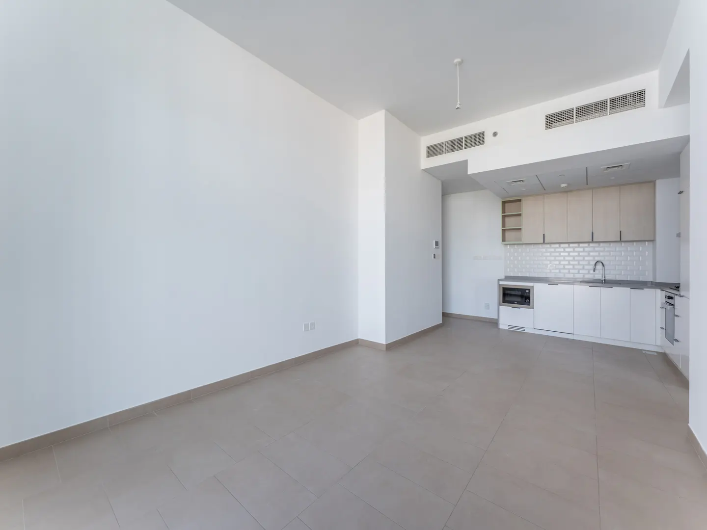 An empty room with white walls and beige tile floors leads to a kitchen with white cabinets and stainless steel appliances.