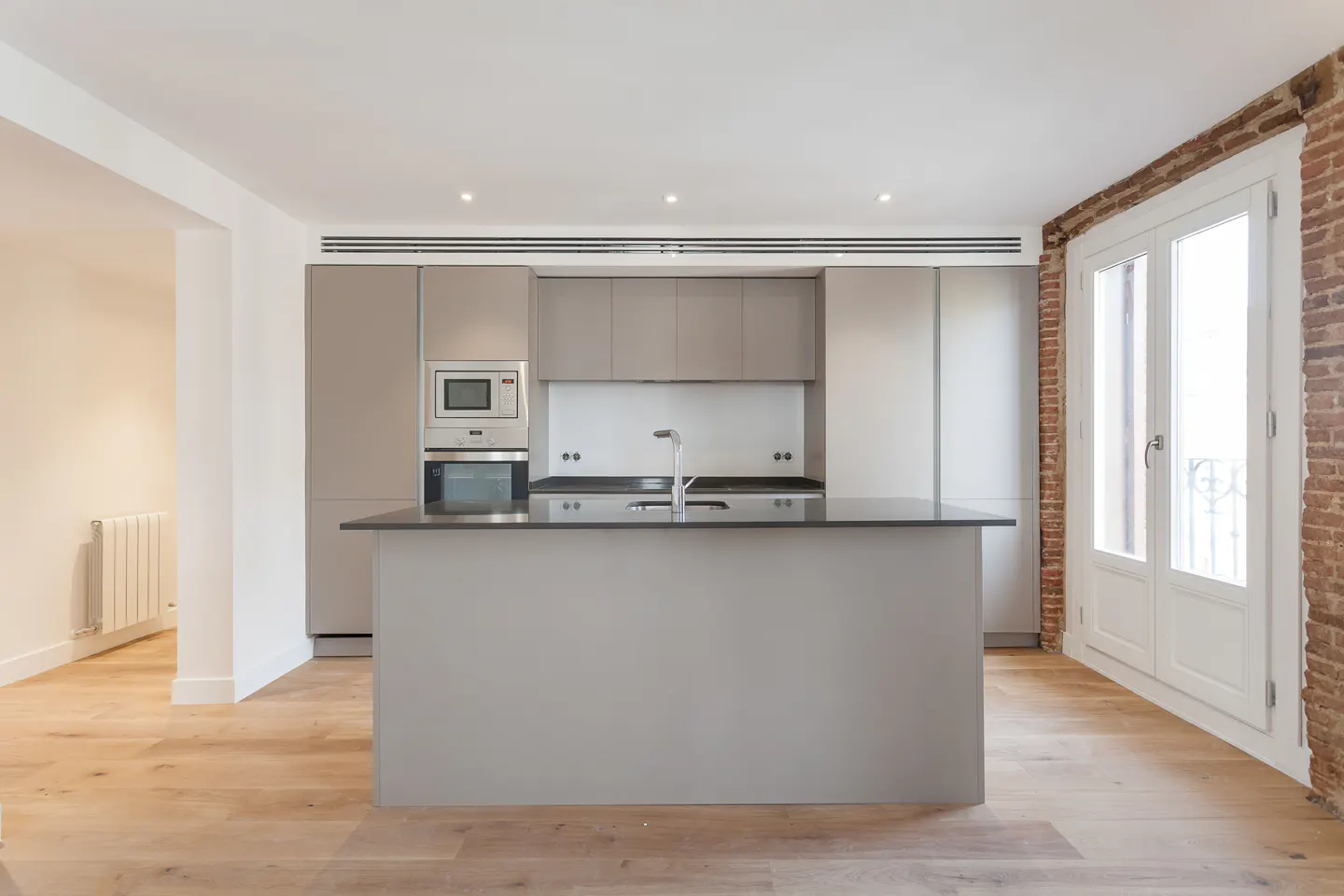 Bright kitchen with gray cabinets, black countertops, and a central island. Hardwood floors and exposed brick wall add texture. White double doors lead to a balcony.