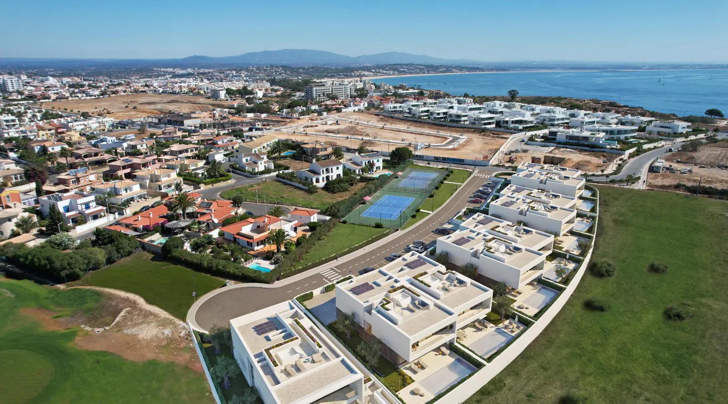 Aerial view of a coastal community with white modern homes, tennis courts, and ocean backdrop under a clear blue sky.