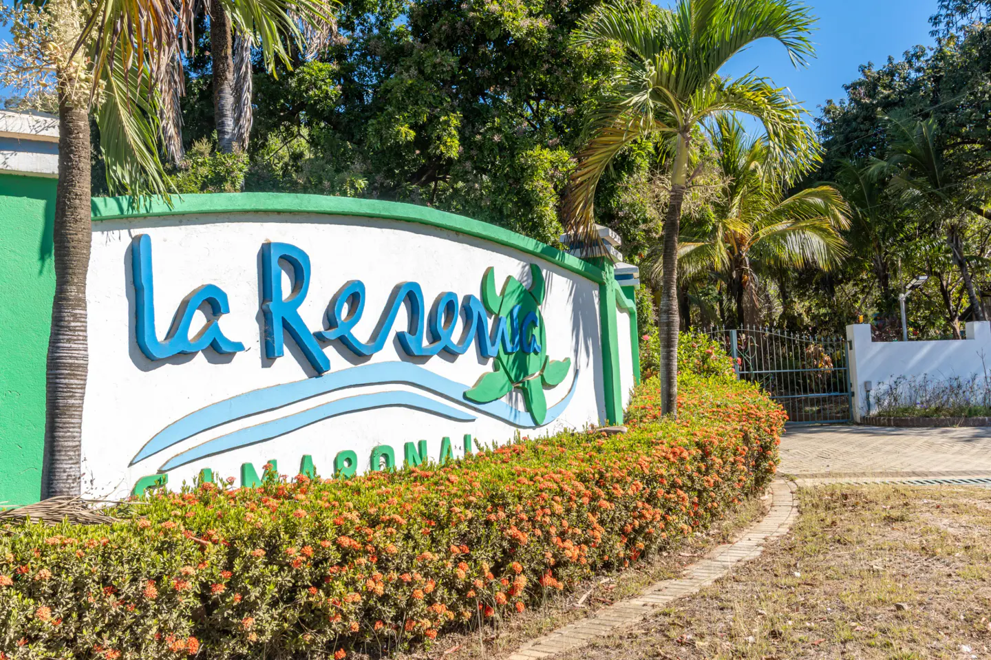 Entrance sign to "La Reserva Camaronal" with blue lettering, green trim, and orange flowers. Palm trees and a gate are in the background.