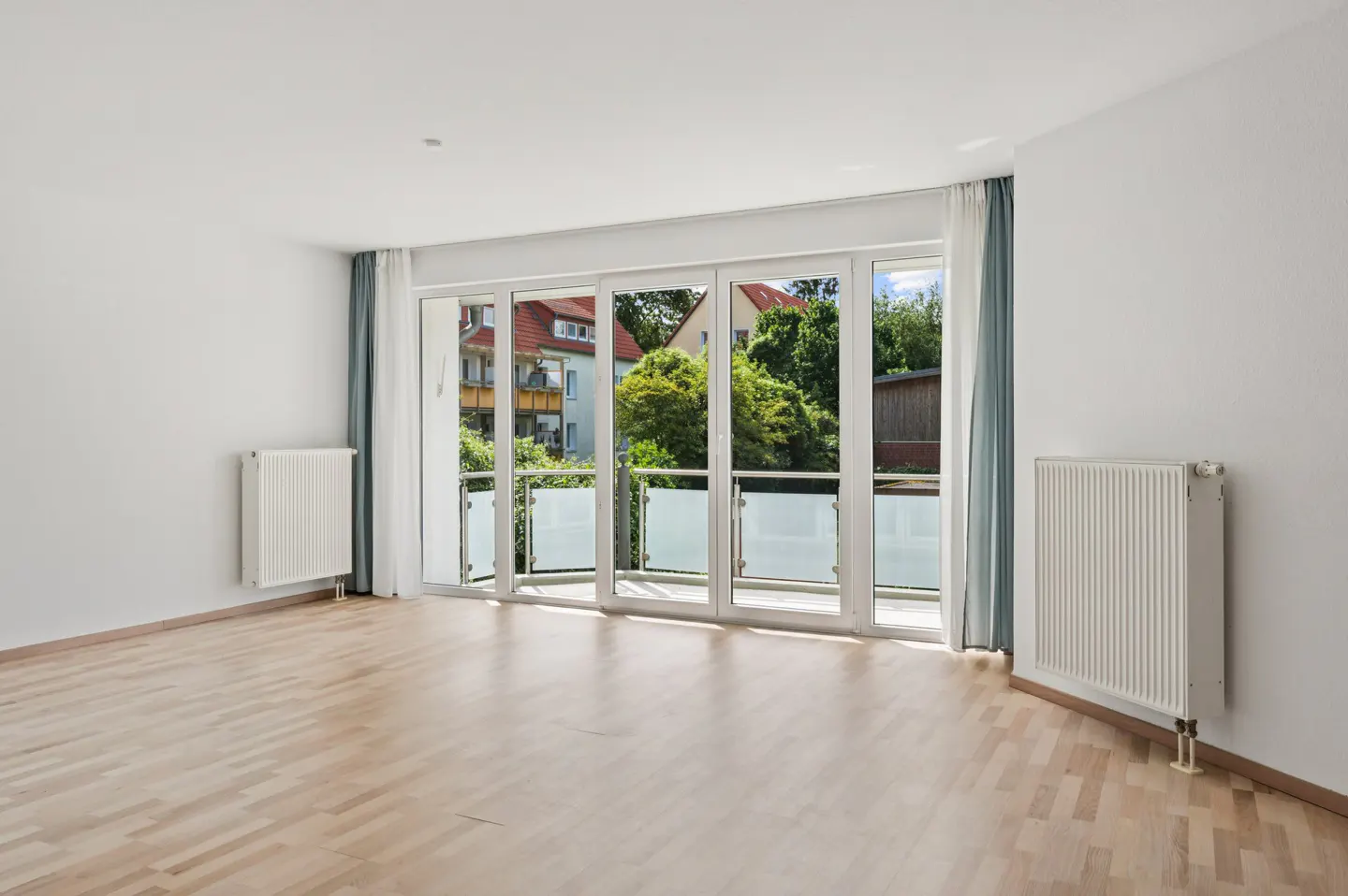 Bright, empty room with light wood floors, white walls, two radiators, and large glass doors opening to a balcony.