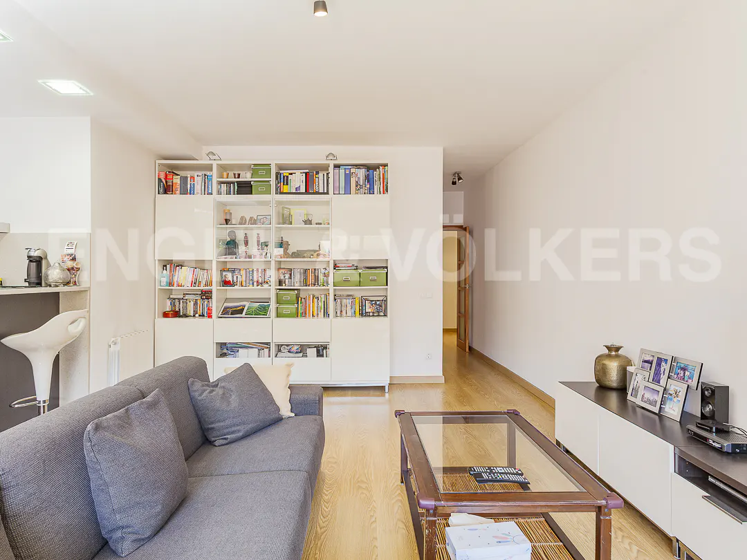 Living room with gray sofa, glass coffee table, and white bookcase filled with books and decor. Hardwood floors and white walls.