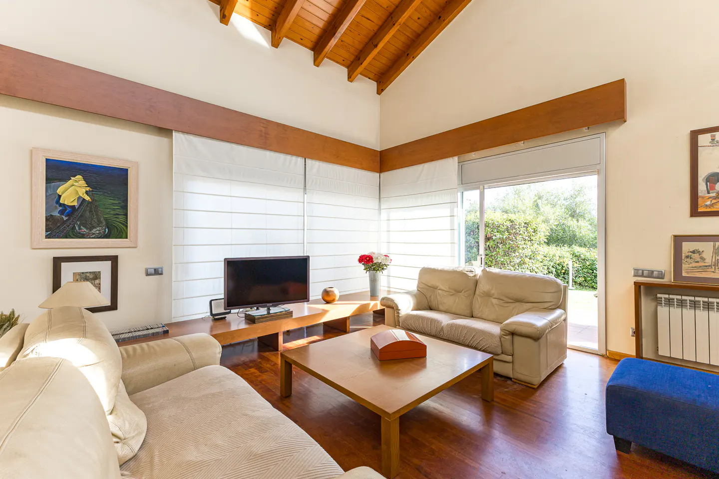 A living room with beige sofas, a wooden coffee table, and a TV. The room has a high ceiling with wooden beams and a large window.