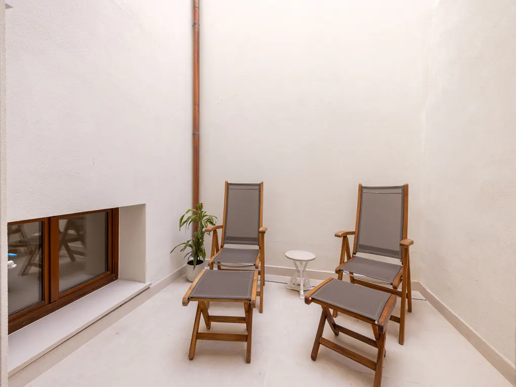 A bright, minimalist patio with two wooden chairs, footstools, and a small white table. A window and a potted plant add to the serene setting.