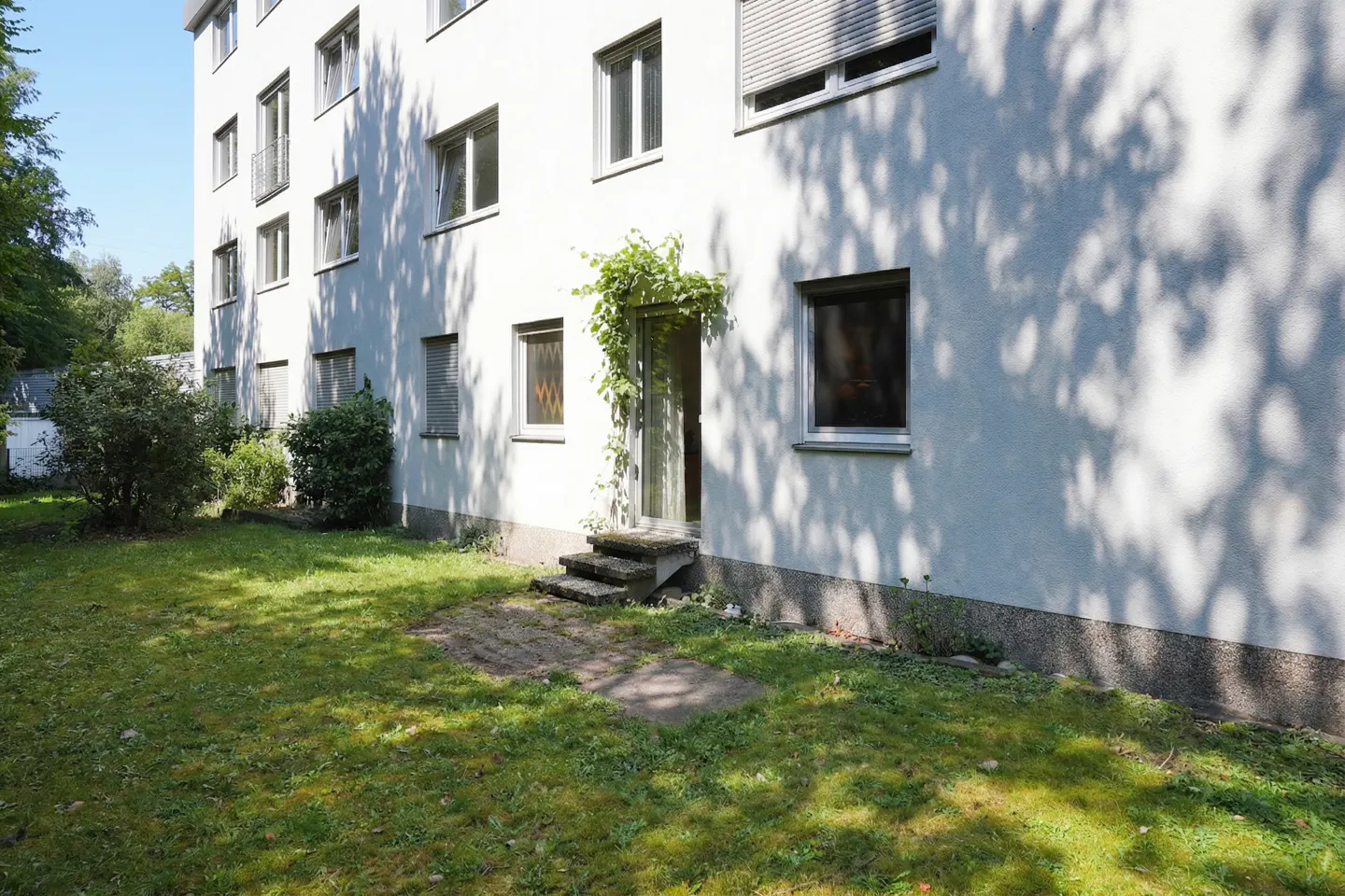 Exterior view of a white apartment building with a green lawn, steps, and a vine-covered entrance.