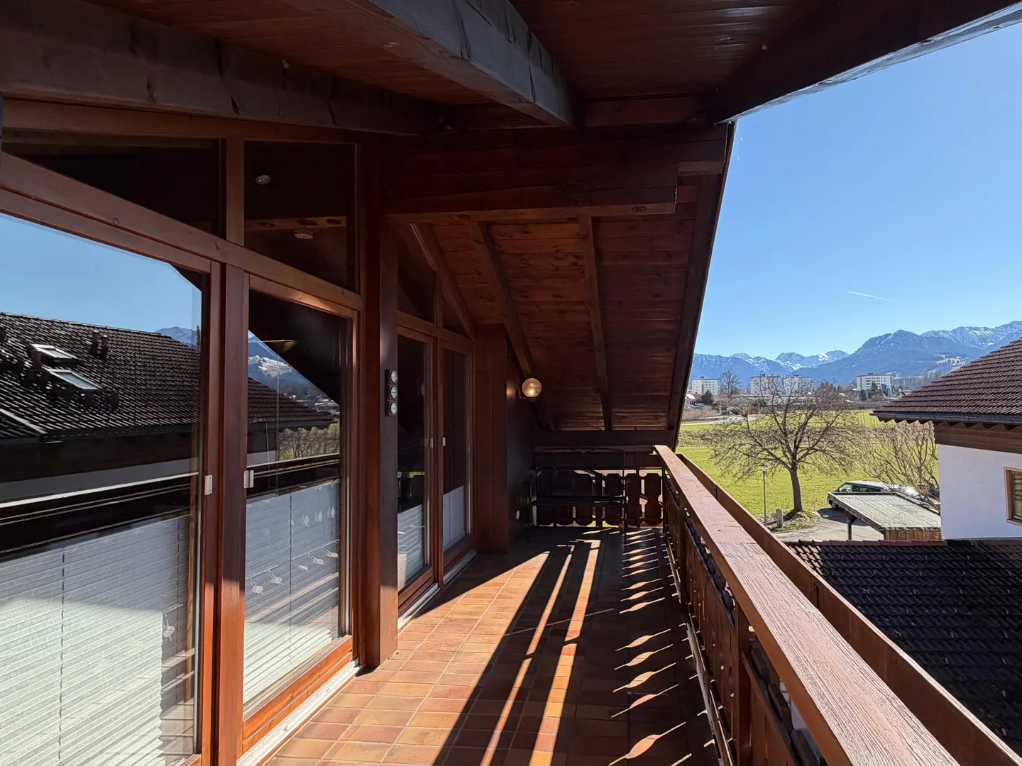Balcony view with brown wood ceiling, brick floor, and metal railing. Mountains and blue sky are visible in the background.