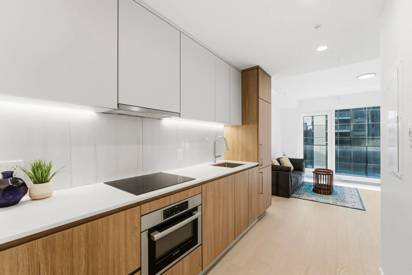 Modern kitchen with white cabinets, wood-grain lower cabinets, stainless steel oven, and a view of the living room.