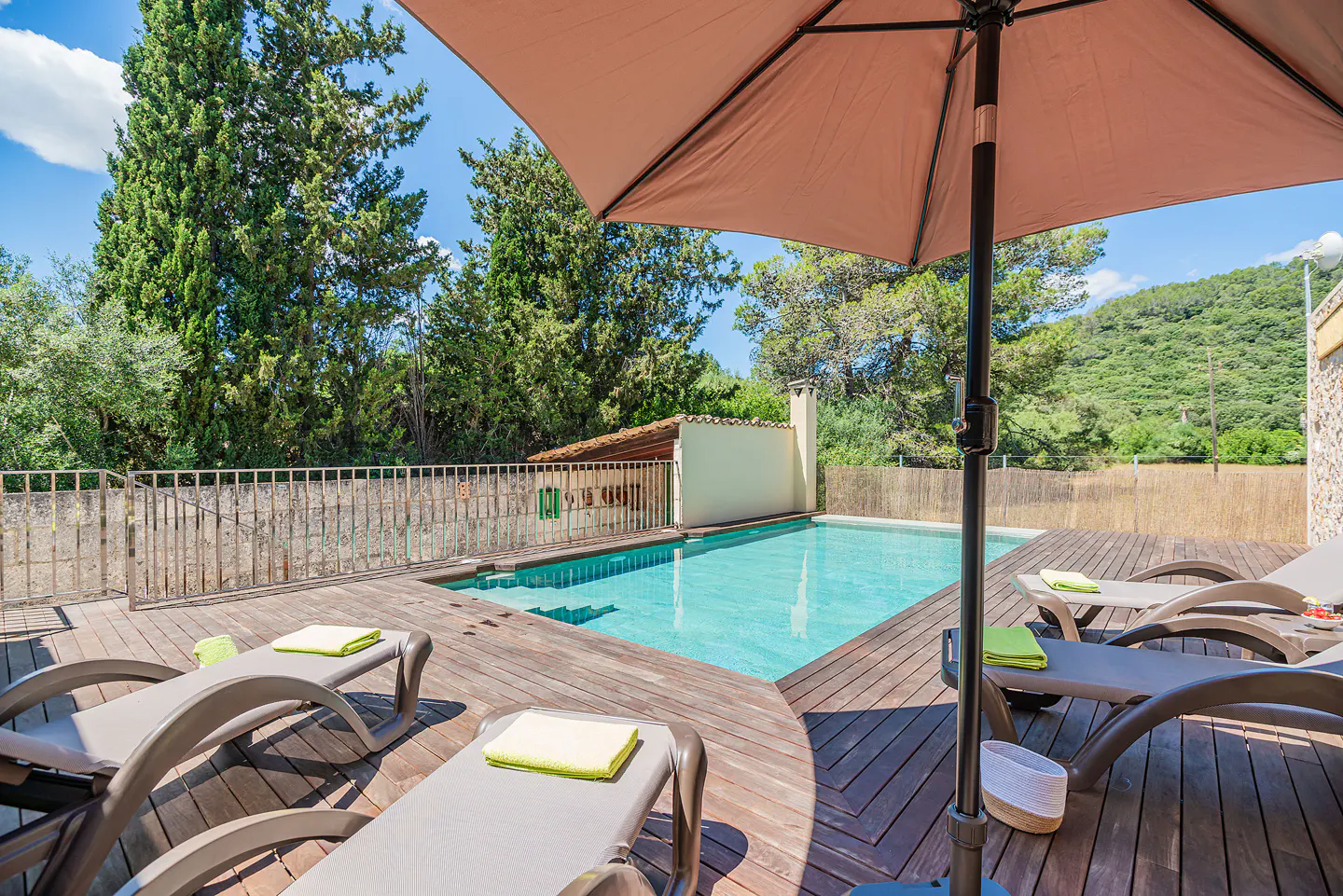 Outdoor pool with turquoise water, surrounded by a wooden deck, lounge chairs with green towels, and a tan umbrella.