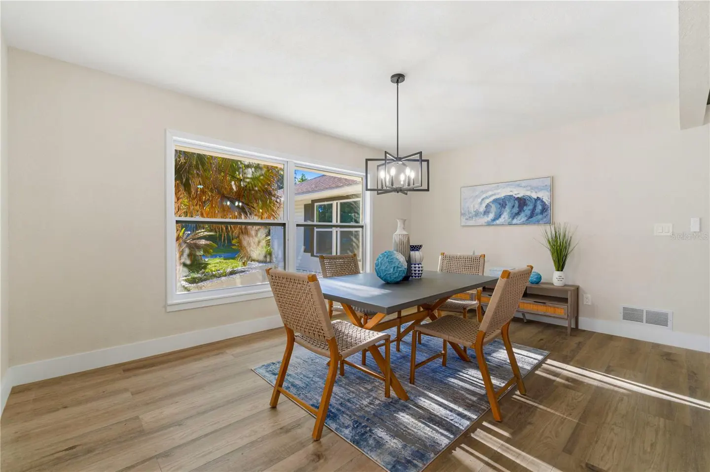 Bright dining room with wood floors, a gray table, and woven chairs on a blue rug. A window overlooks a green yard.