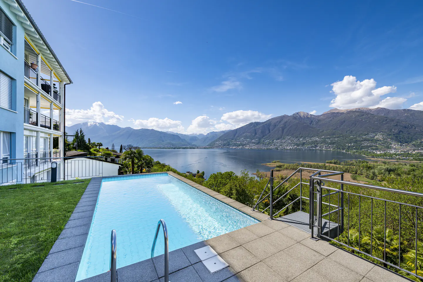 Outdoor pool with metal ladder and patio overlooking a lake and mountains under a blue sky.