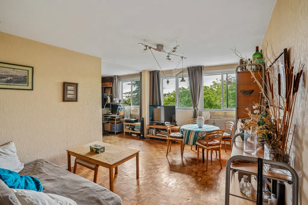 A bright living room with parquet floors, a sofa, a table, and a dining area with a view of green trees.