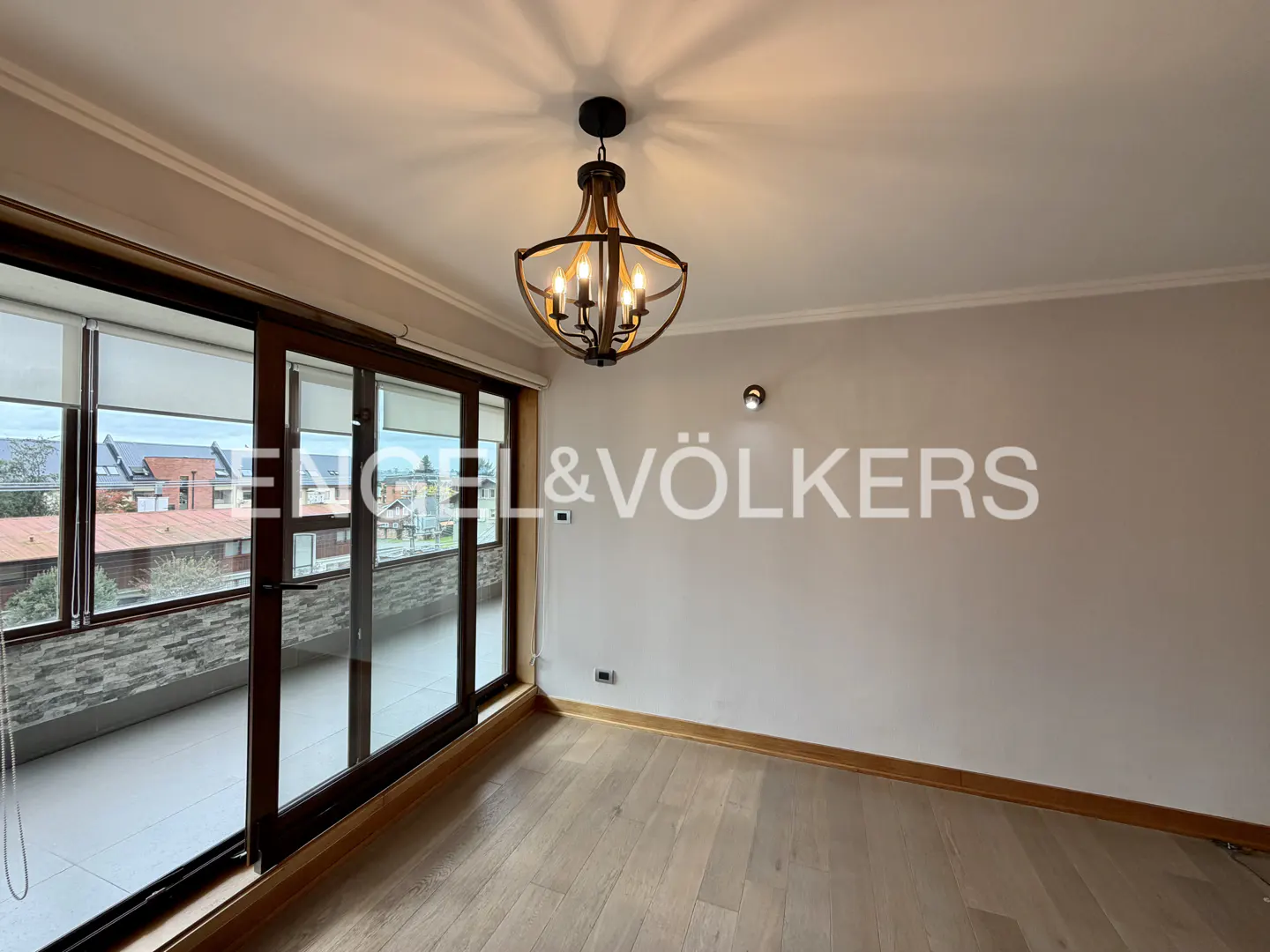 Empty room with light wood floors, white walls, and a chandelier. Sliding glass doors lead to a balcony with a view of houses.