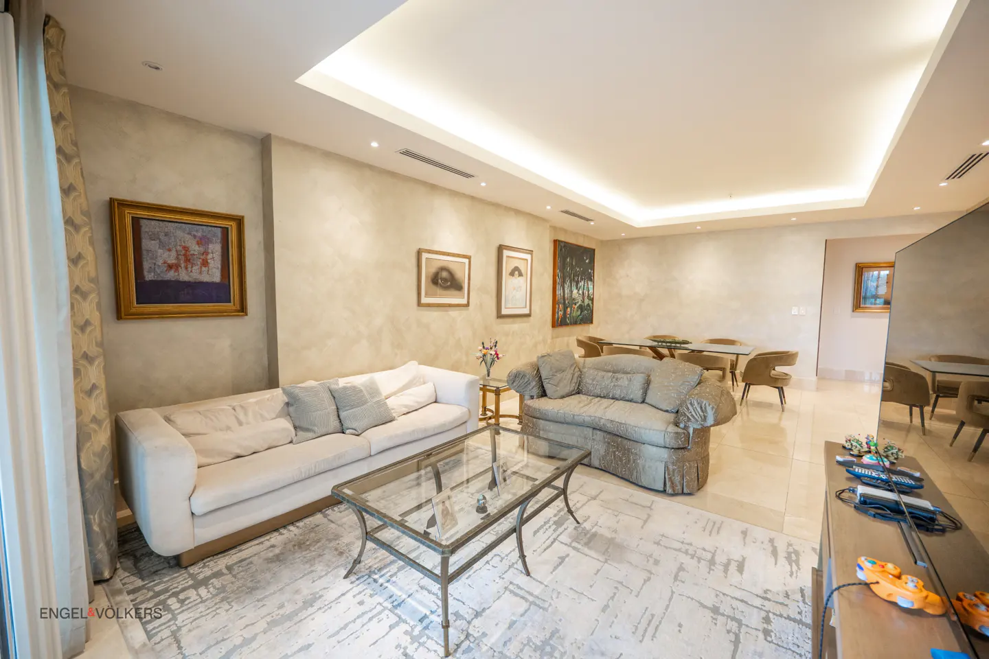 A living room with a white sofa, gray sofa, glass table, and dining table with chairs. Art hangs on the beige walls.