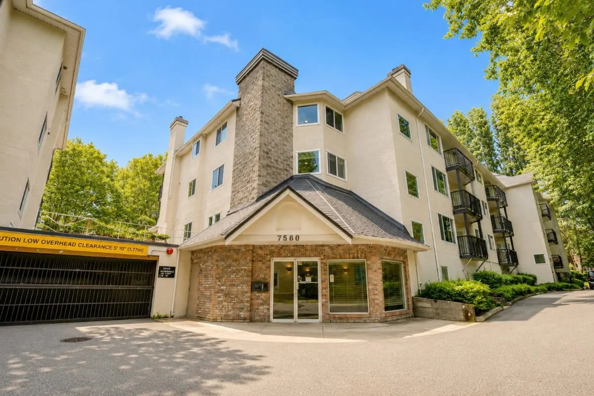 Exterior view of a multi-story beige apartment building with a brick entrance, stone chimney, and underground parking.