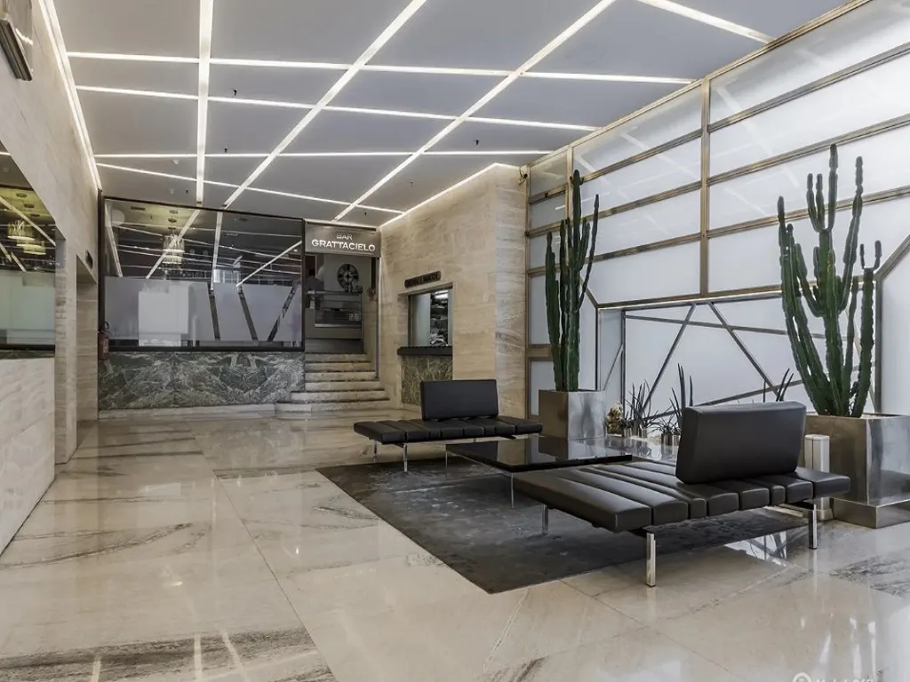 Modern lobby with marble floors, black leather seating, and tall cacti. Geometric ceiling lights add a contemporary touch.