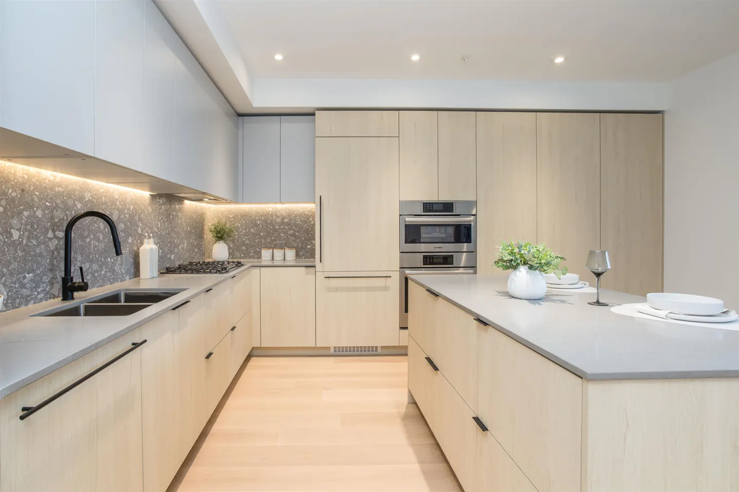 A modern kitchen with light wood cabinets, gray countertops, and stainless steel appliances. An island with dishes and a wine glass sits in the center.