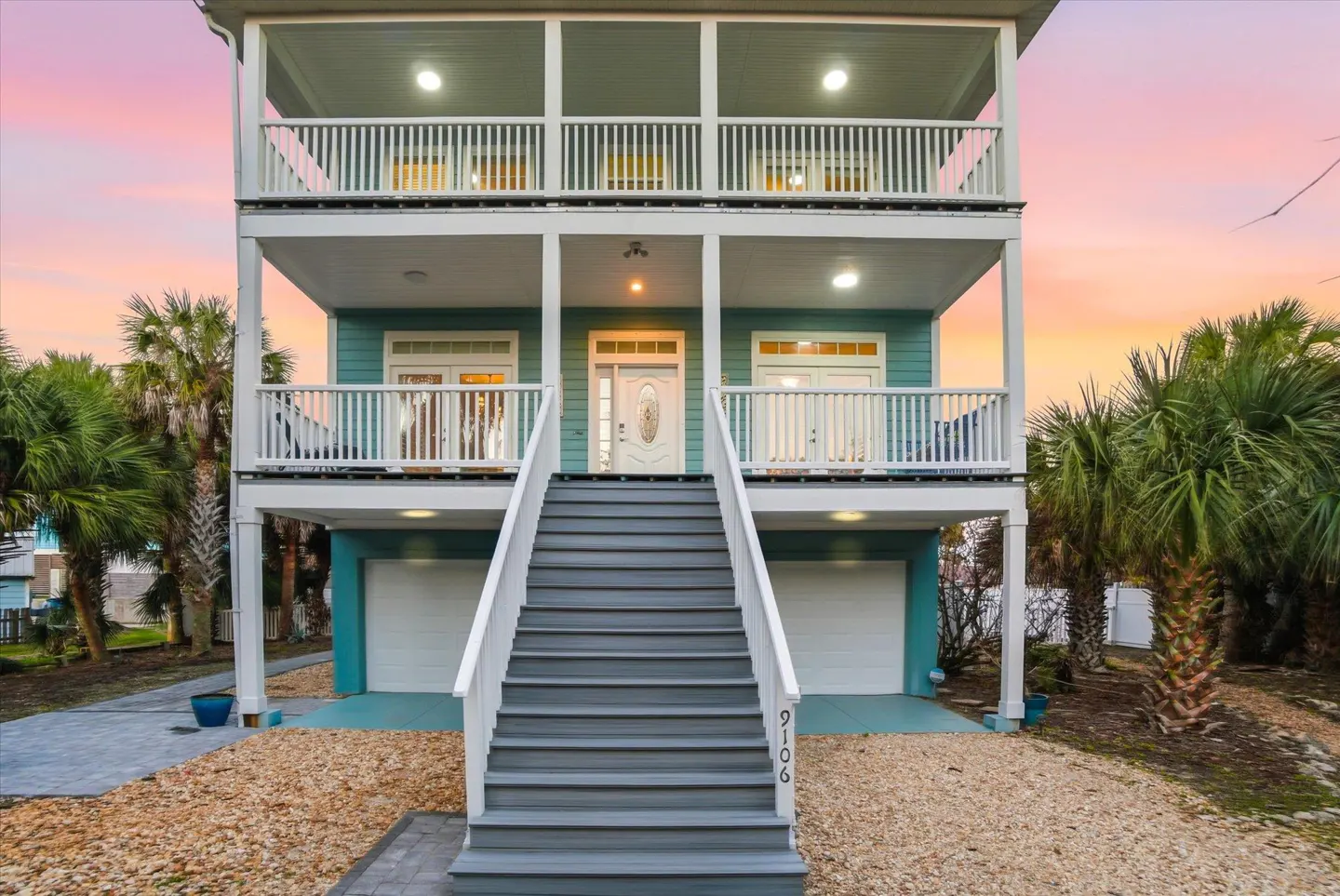 Two-story teal house with white trim, two balconies, and gray stairs leading to the front door. Palm trees surround the house.