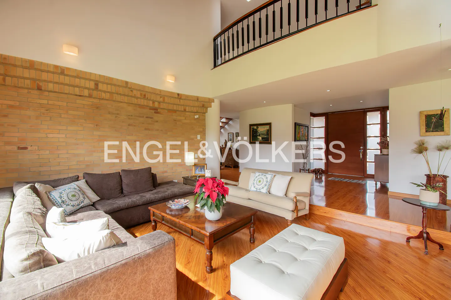 Living room with brown sofas, wood floors, brick wall, and a balcony overlooking the space. A white ottoman sits in front of a wood coffee table.