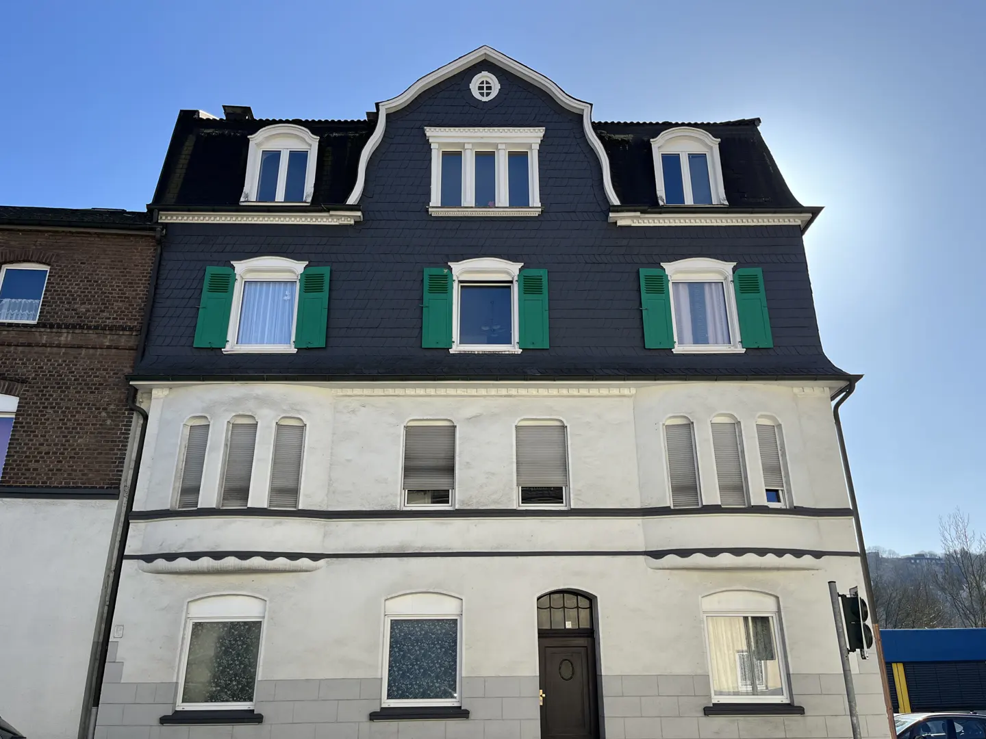 Three-story building with a dark roof, green shutters, and white walls under a blue sky.