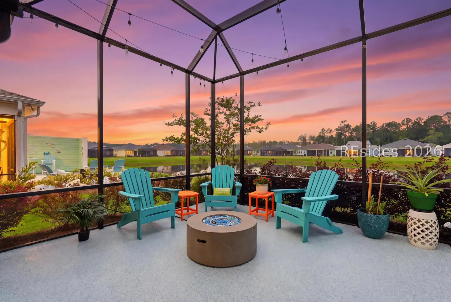 Screened patio with turquoise Adirondack chairs, orange side tables, and a fire pit. Sunset sky and pond in the background.