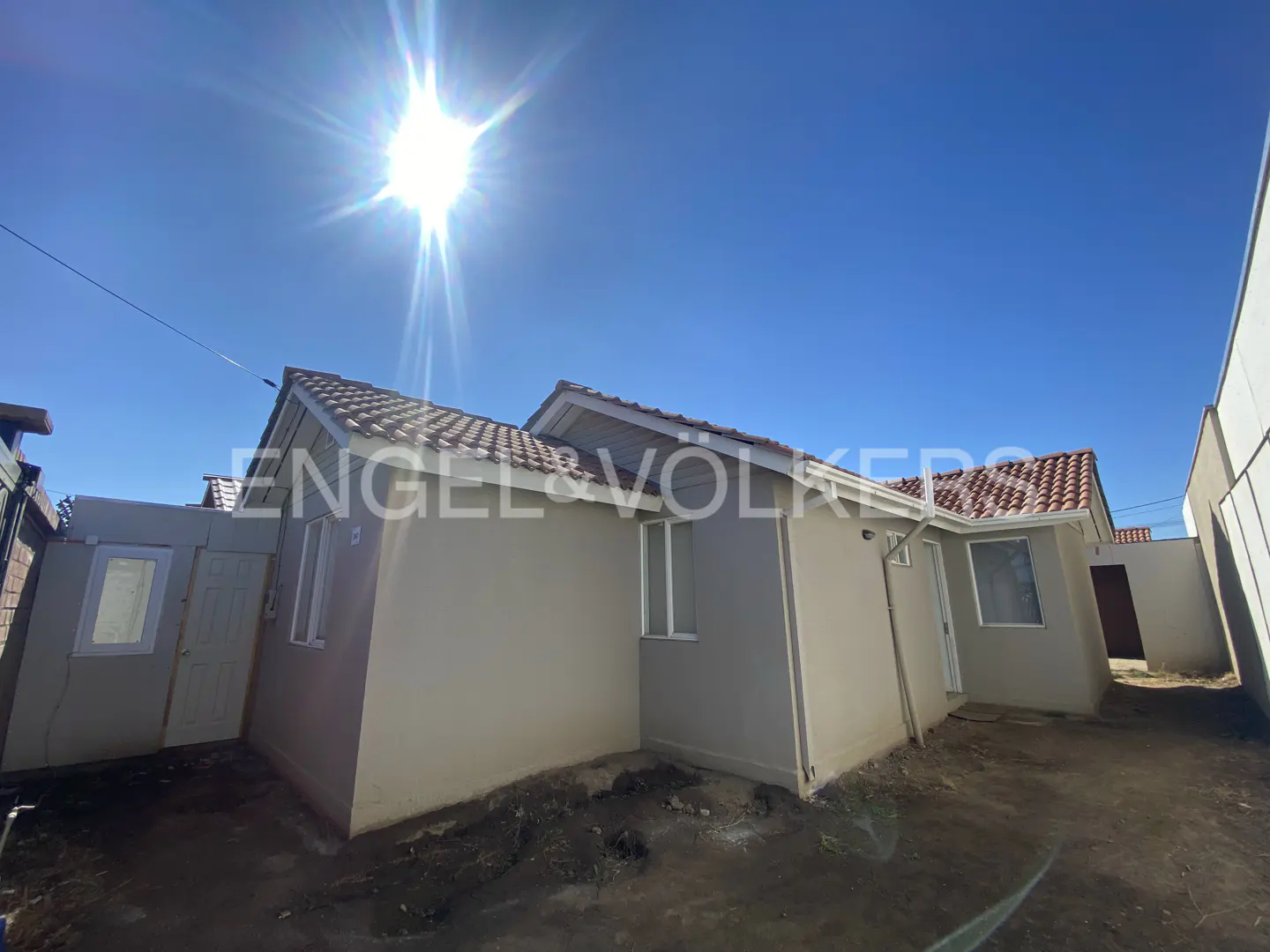 Exterior view of a beige single-story house with a red tile roof under a bright blue sky. The Engel & Völkers logo is visible on the side of the house.