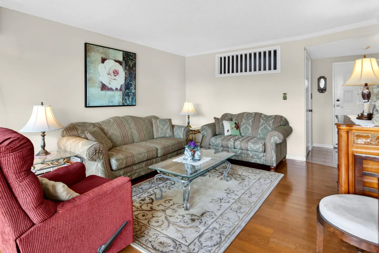 Living room with two patterned sofas, a red recliner, and a glass coffee table on a patterned rug. A rose painting hangs on the wall.