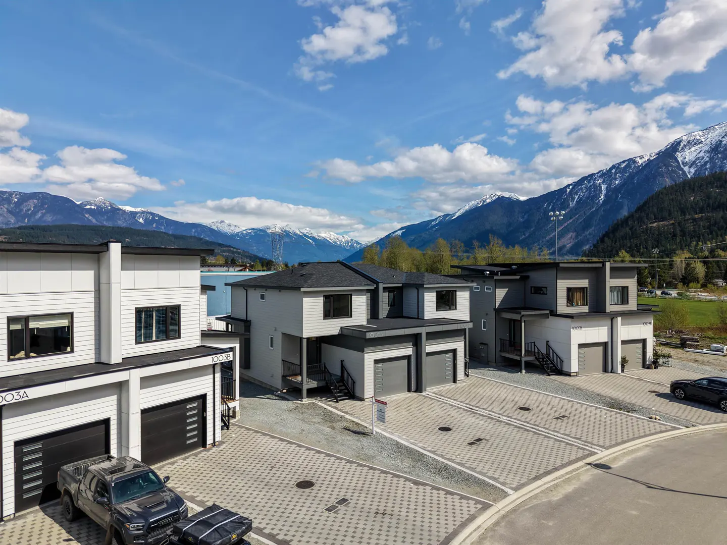 Three modern townhouses with gray and white exteriors, black garage doors, and mountain views under a blue sky.