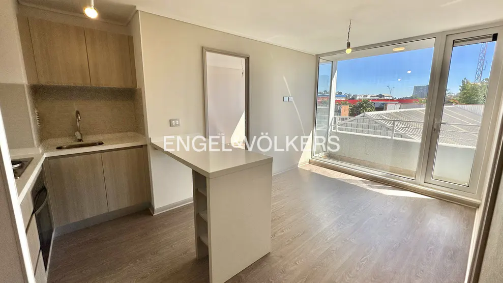 A bright, modern apartment interior with wood floors, a kitchen area with light wood cabinets, and a white island. A large window shows a balcony and city view.