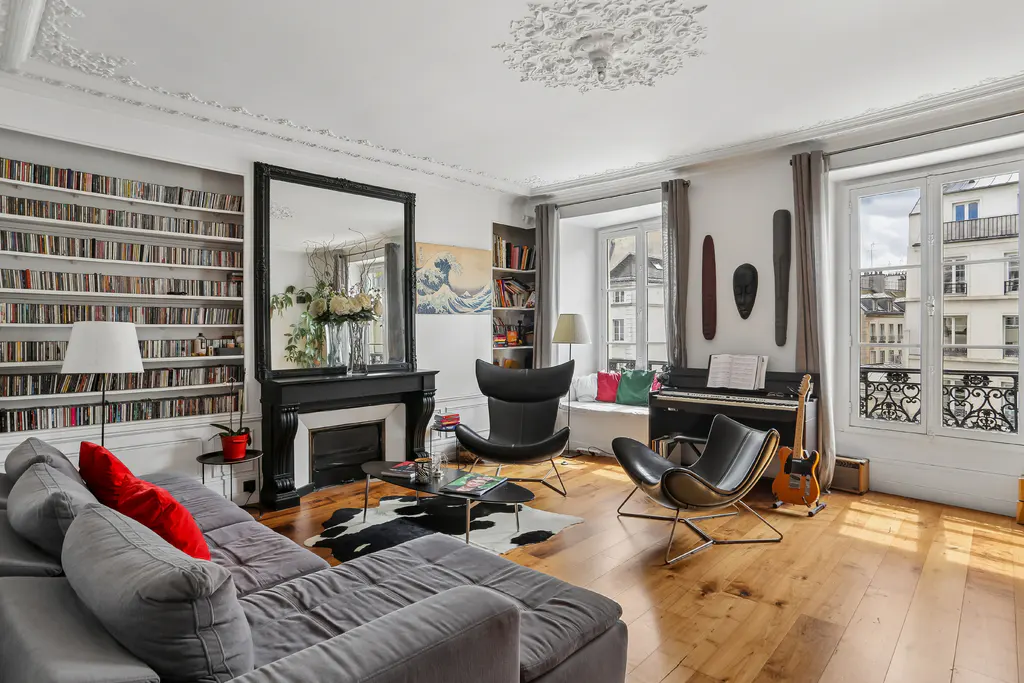 Bright living room with hardwood floors, gray sectional sofa with red pillows, black fireplace, piano, and large windows.