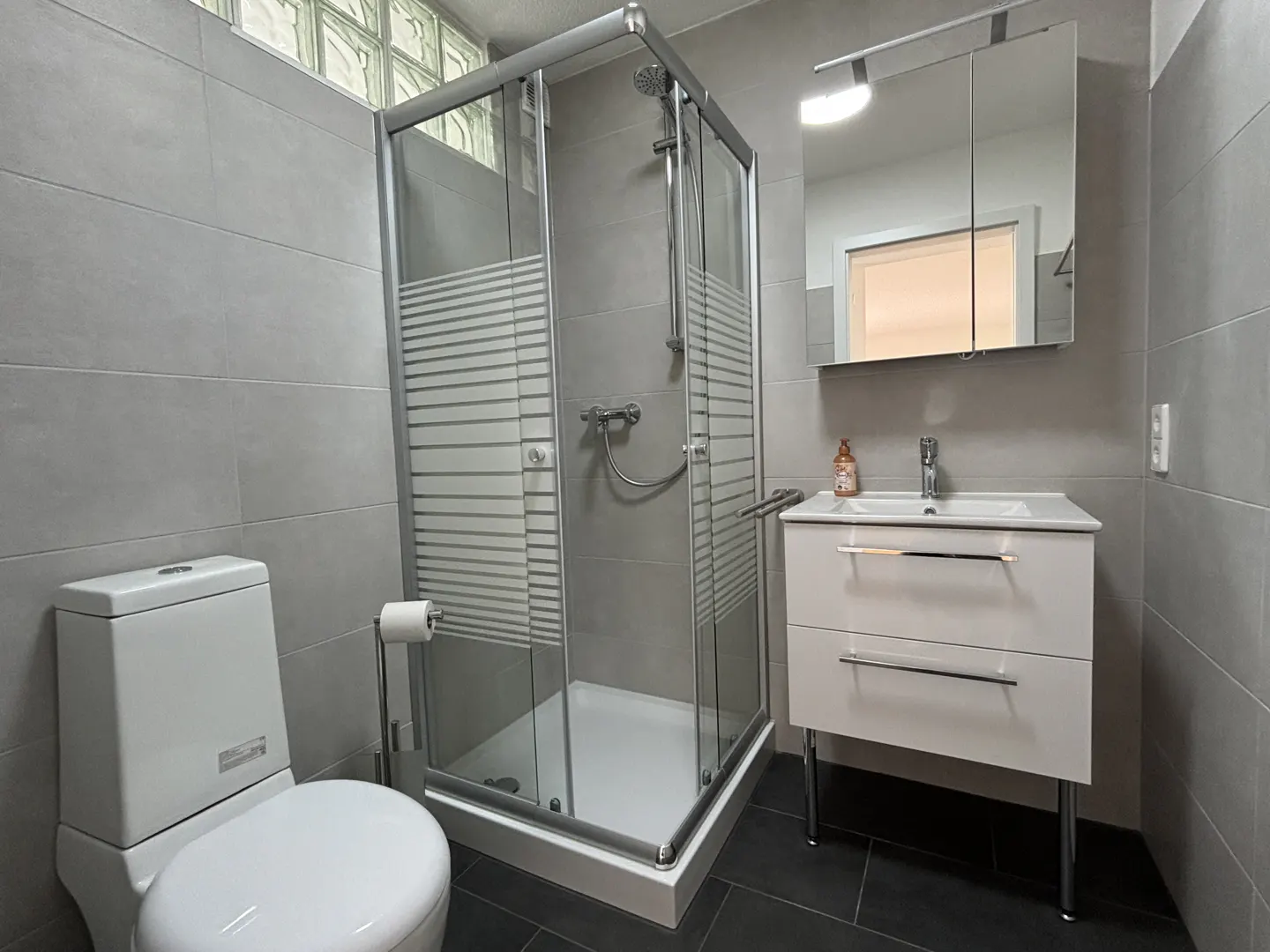 Modern bathroom with gray tiled walls, dark floor, glass shower, white toilet, and white vanity with a mirror.