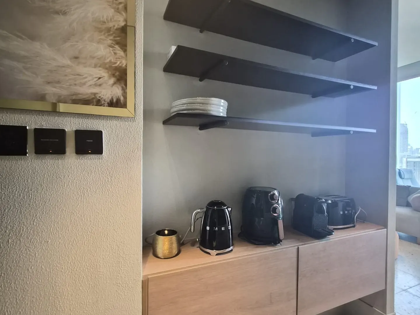 Modern kitchen nook with light wood cabinets, black appliances, floating shelves, and a textured wall with gold-framed art.