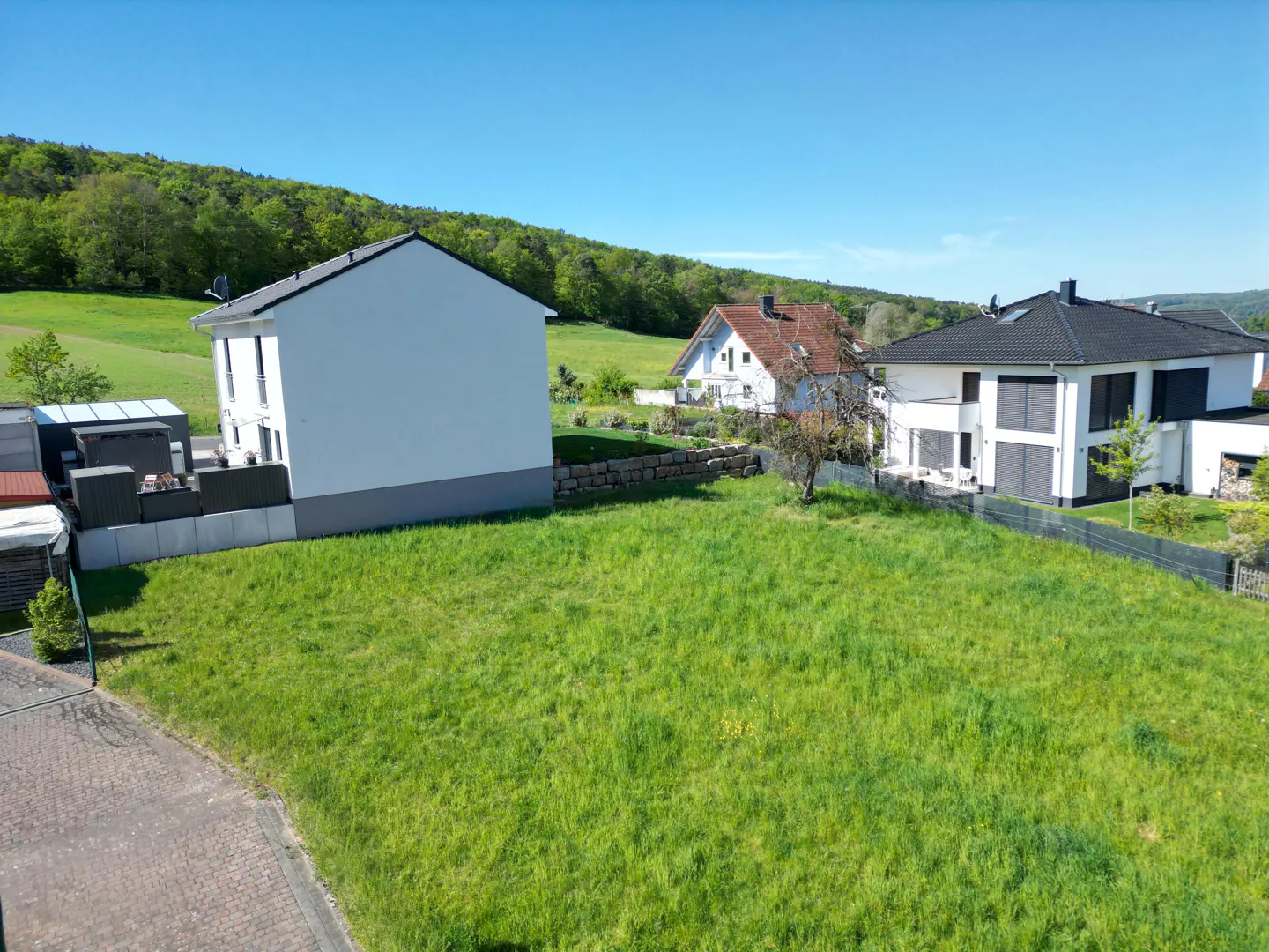 A grassy lot with modern white houses under a blue sky, surrounded by green trees.