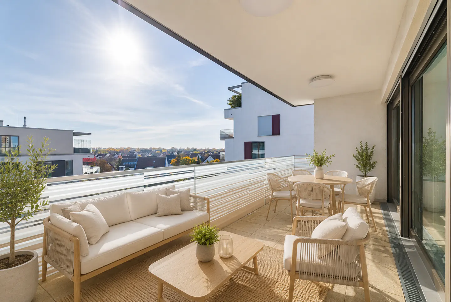 A sunlit balcony with a sofa, chairs, and table. A view of buildings and trees is in the background.