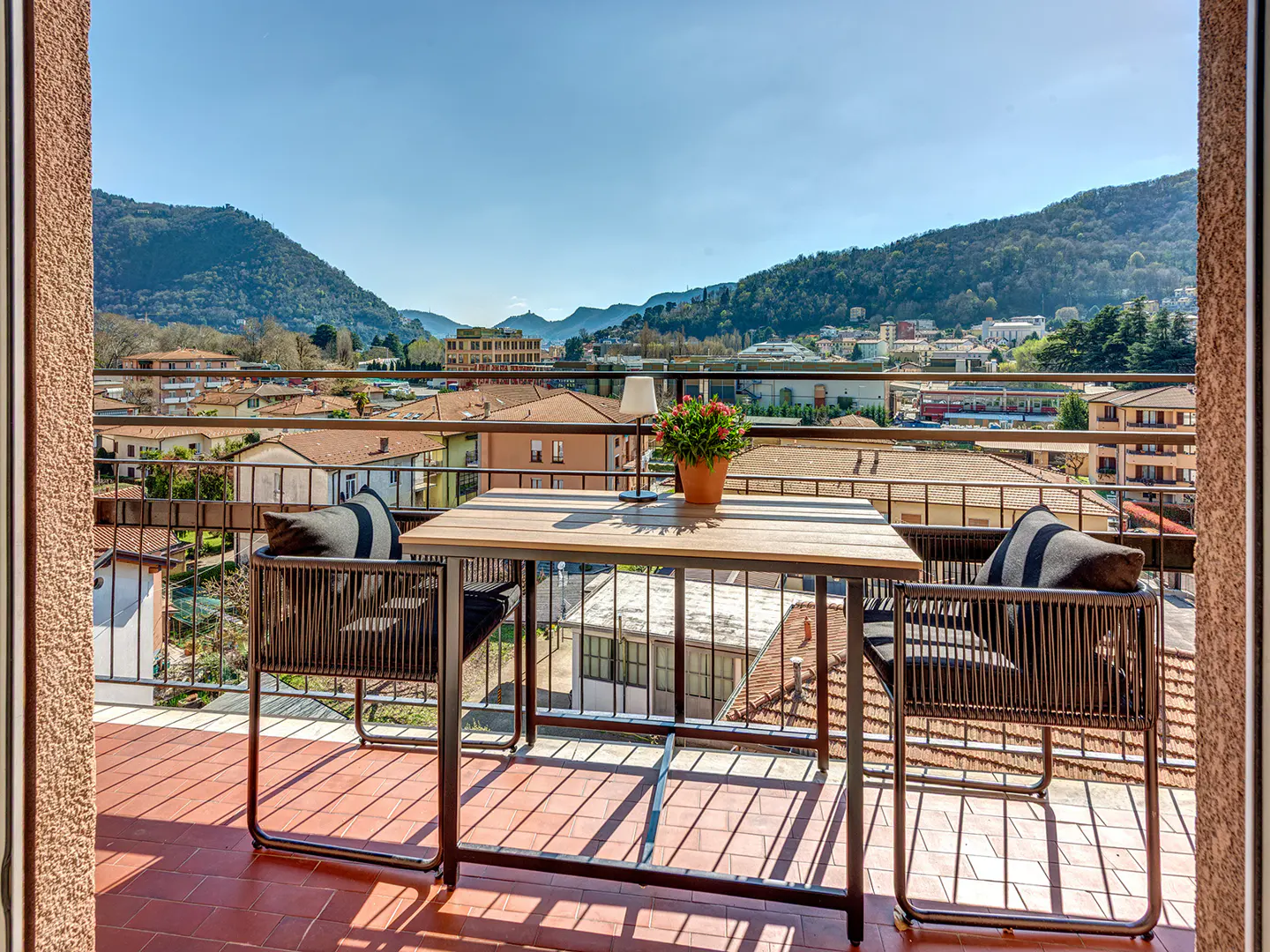 Balcony view with table, chairs, and city backdrop. The table has a lamp and potted plant. The chairs have striped pillows.