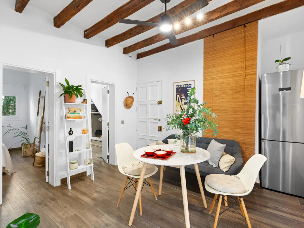 Bright living space with wood floors, white walls, and exposed beams. A round table with chairs sits near a gray sofa and stainless steel refrigerator.