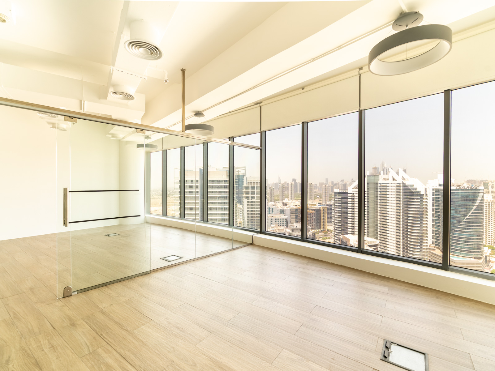 Bright, empty office space with light wood floors, glass partitions, and floor-to-ceiling windows overlooking a city skyline.