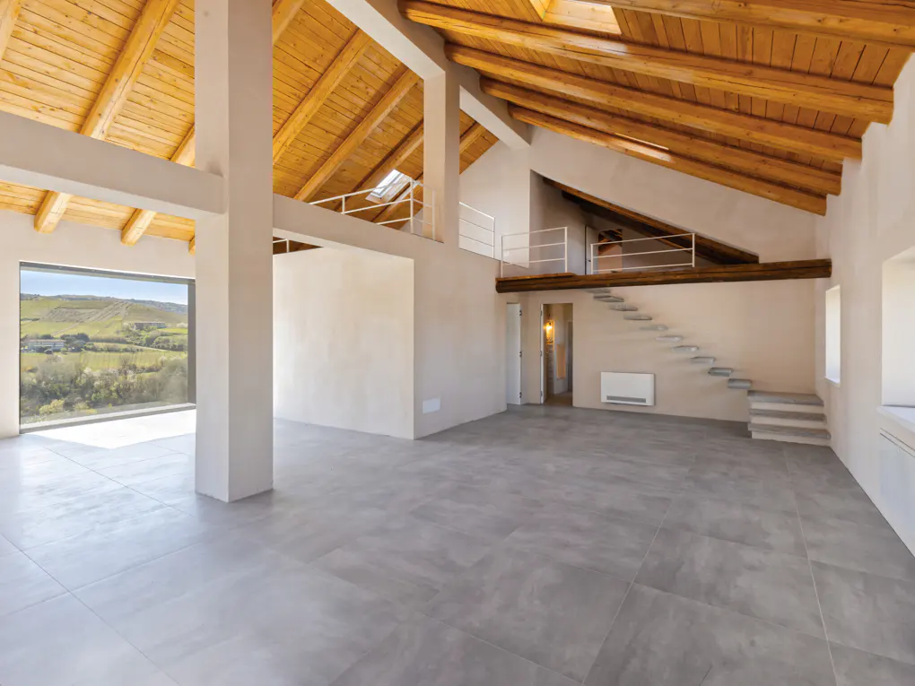 Bright, empty loft with gray tile floors, white walls, and a wooden beamed ceiling. A staircase leads to a small loft area. A large window shows a green landscape.