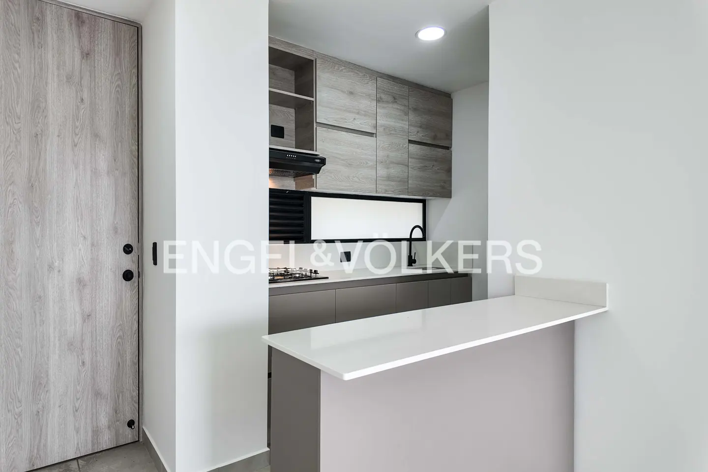 A modern kitchen with gray cabinets, a white countertop, and a light wood door. The kitchen features a gas stove, a black faucet, and a window.