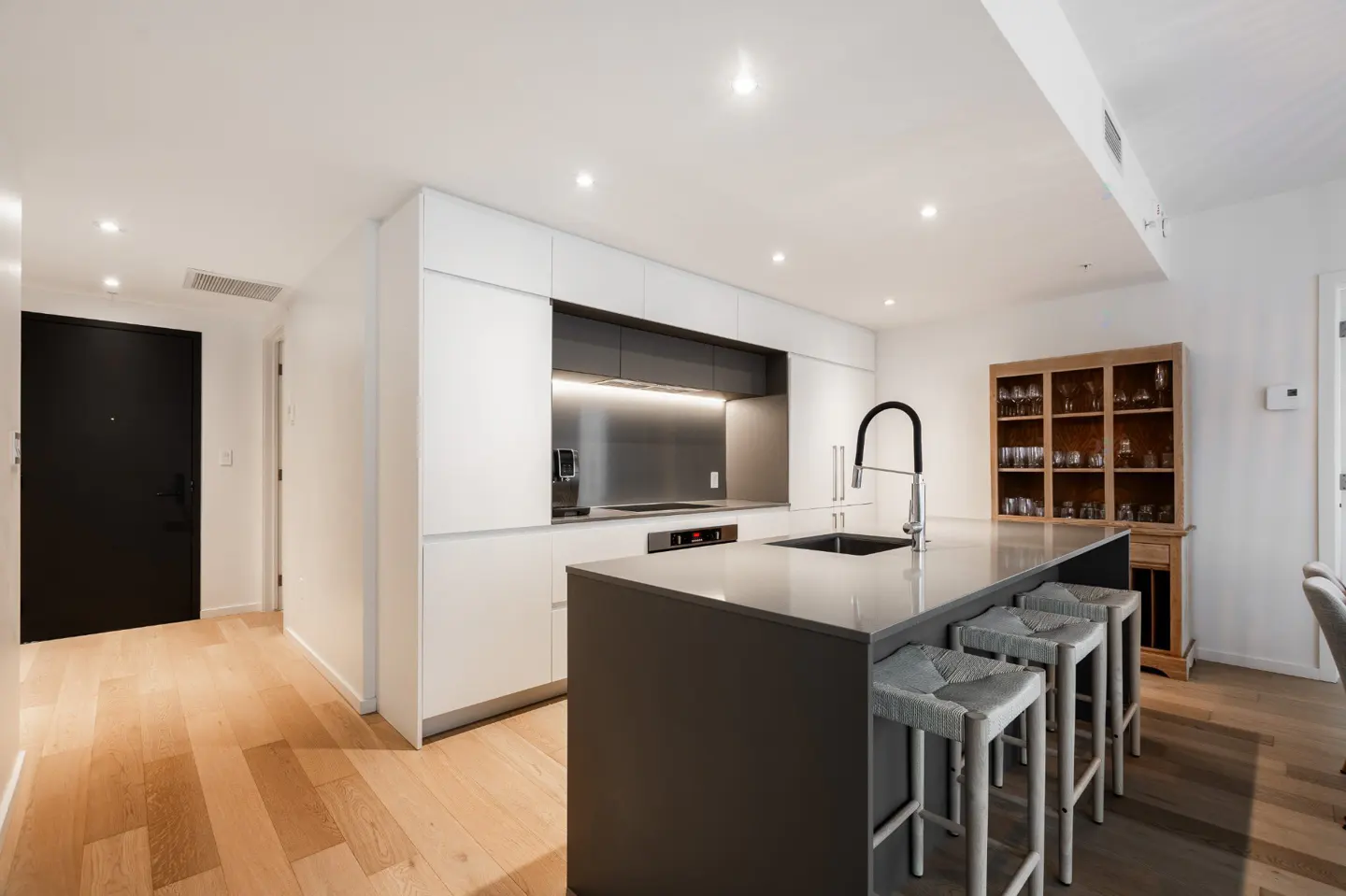 Bright, modern kitchen with a gray island, white cabinets, and wooden floors. Three woven stools sit at the island. A black door is visible in the background.