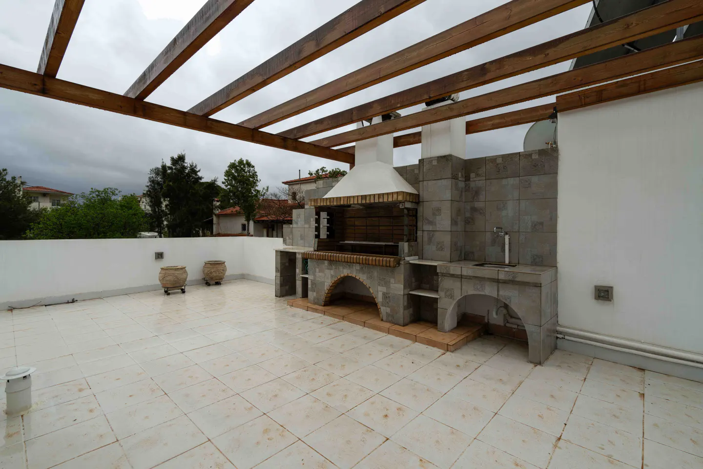 Outdoor patio with a built-in stone grill and sink under a wooden pergola. Two decorative pots sit against a white wall.