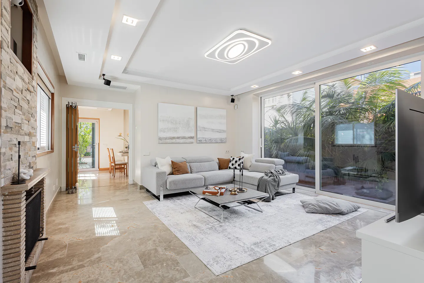 Bright living room with a gray sofa, coffee table, and large windows overlooking a lush garden. A stone fireplace adds texture.