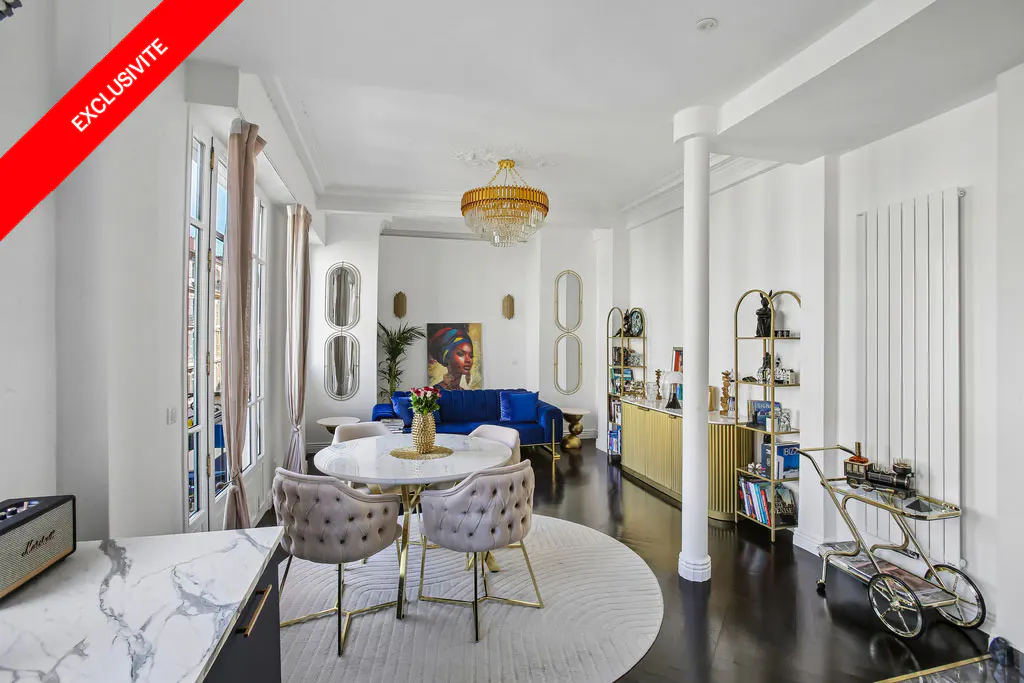 Bright living room with white walls, dark wood floors, and a round table with beige chairs on a white rug. A blue sofa and gold accents add pops of color.