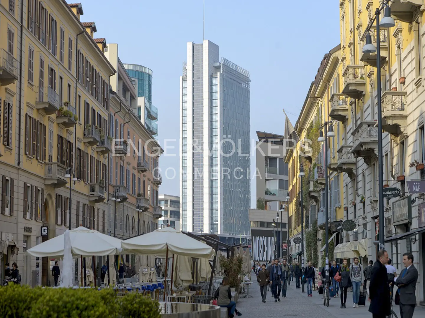 Street view in Milan, Italy, with yellow buildings, shops, and a modern skyscraper in the background. People are walking on the sidewalk.