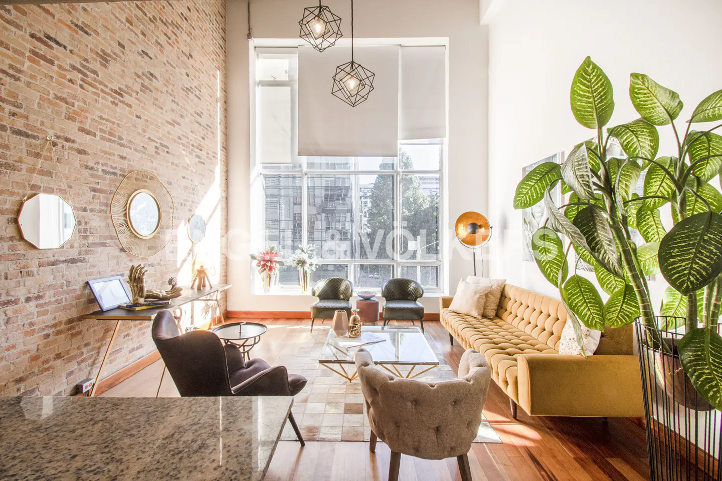 Bright living room with exposed brick wall, hardwood floors, and large windows. A yellow sofa and green plant add color to the space.