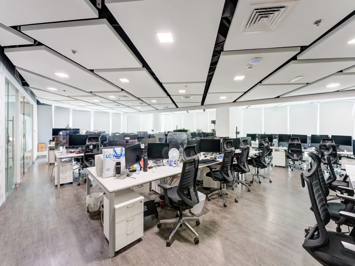 Bright, modern office space with rows of white desks, black chairs, and computer monitors. White ceiling panels and light wood-look floors.