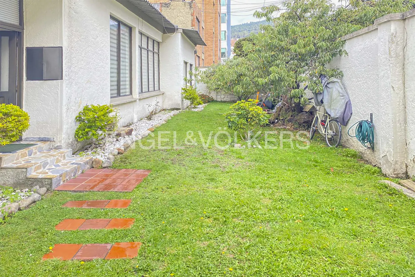 A side yard with a red brick path leading to a white house, green grass, and a bicycle leaning against a white wall.