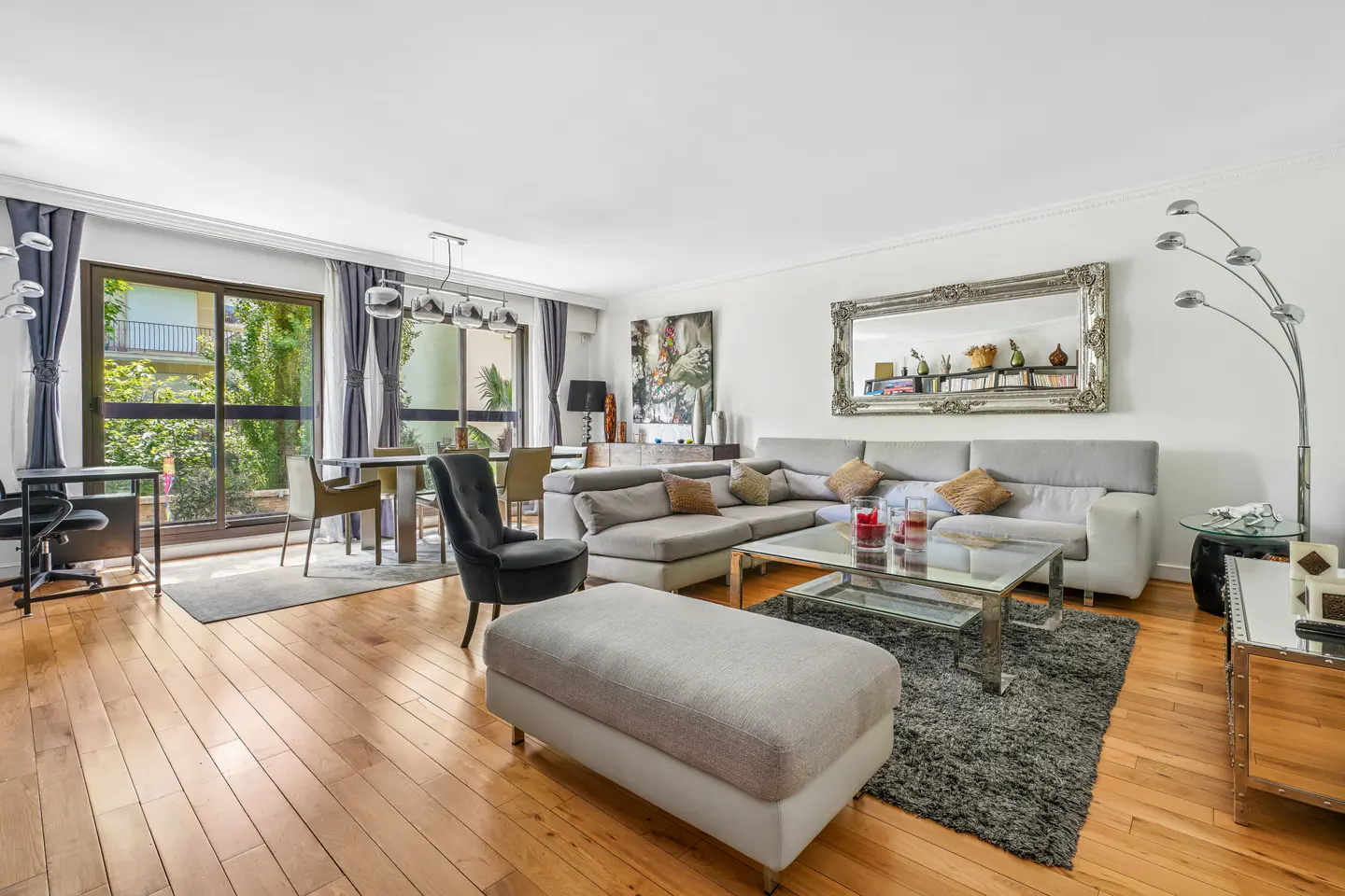 Bright living room with wood floors, a gray sectional sofa, glass coffee table, and large windows with gray curtains.