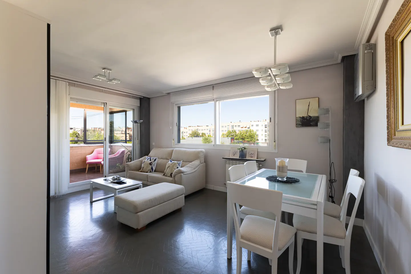 Bright living and dining room with a beige sofa, white dining set, and a balcony featuring pink chairs.