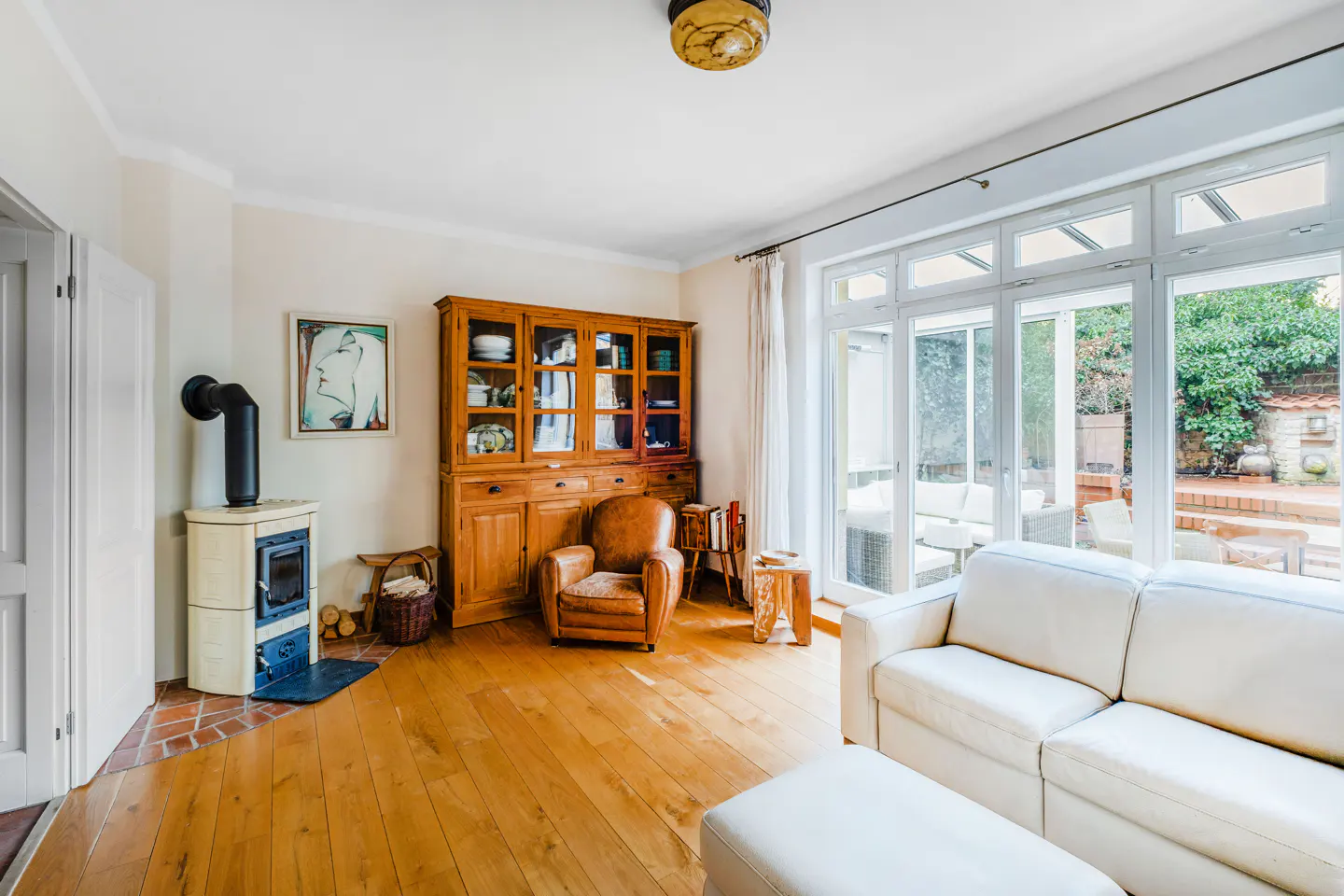 Bright living room with wood floors, white sofa, wood-burning stove, and large windows overlooking a patio.