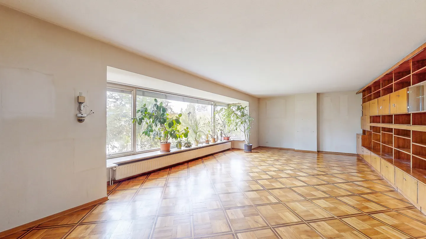 An empty room with parquet floors, a large window with plants, and a built-in wooden shelving unit.